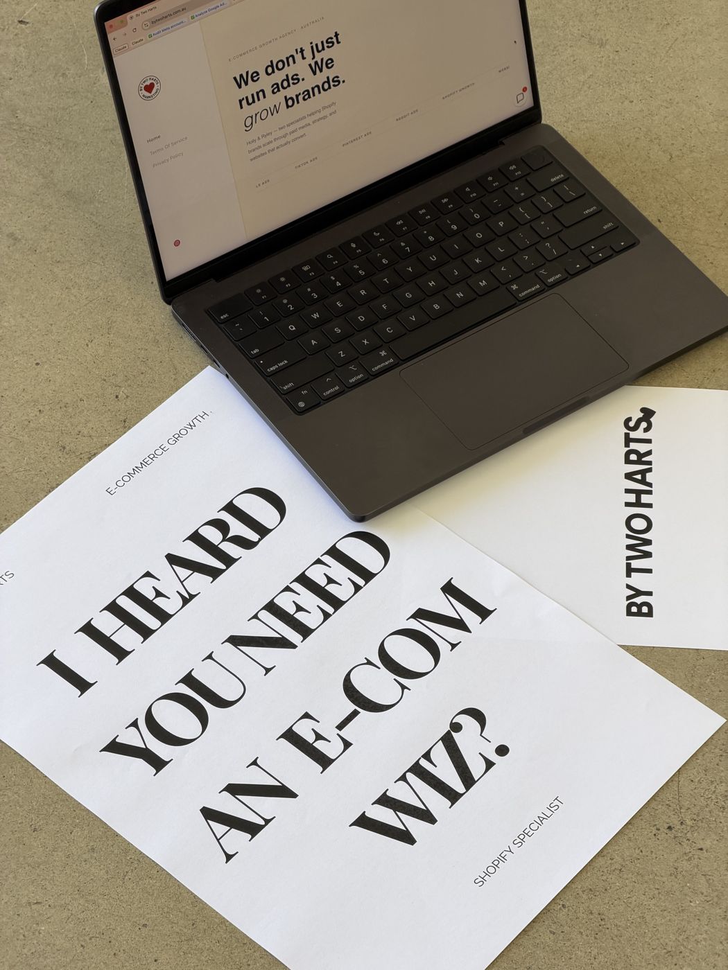 A printed sign on a desk reading I heard you need an e-com wiz, beside a laptop